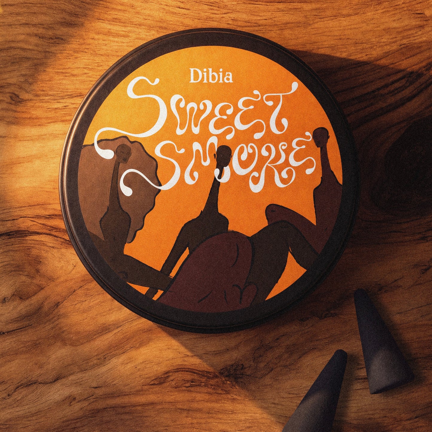 A round black tin labeled 'Dibia Sweet Smoke' sits on a warm wooden surface, with three dark conical shisha tobacco pieces placed nearby. The lid artwork features an orange background with white decorative lettering and dark silhouettes of people smoking.