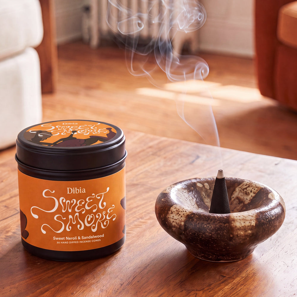 A jar of Dibia Sweet Smoky incense next to a ceramic incense holder with a smoking cone, set on a wooden table in a warm, softly blurred living room.