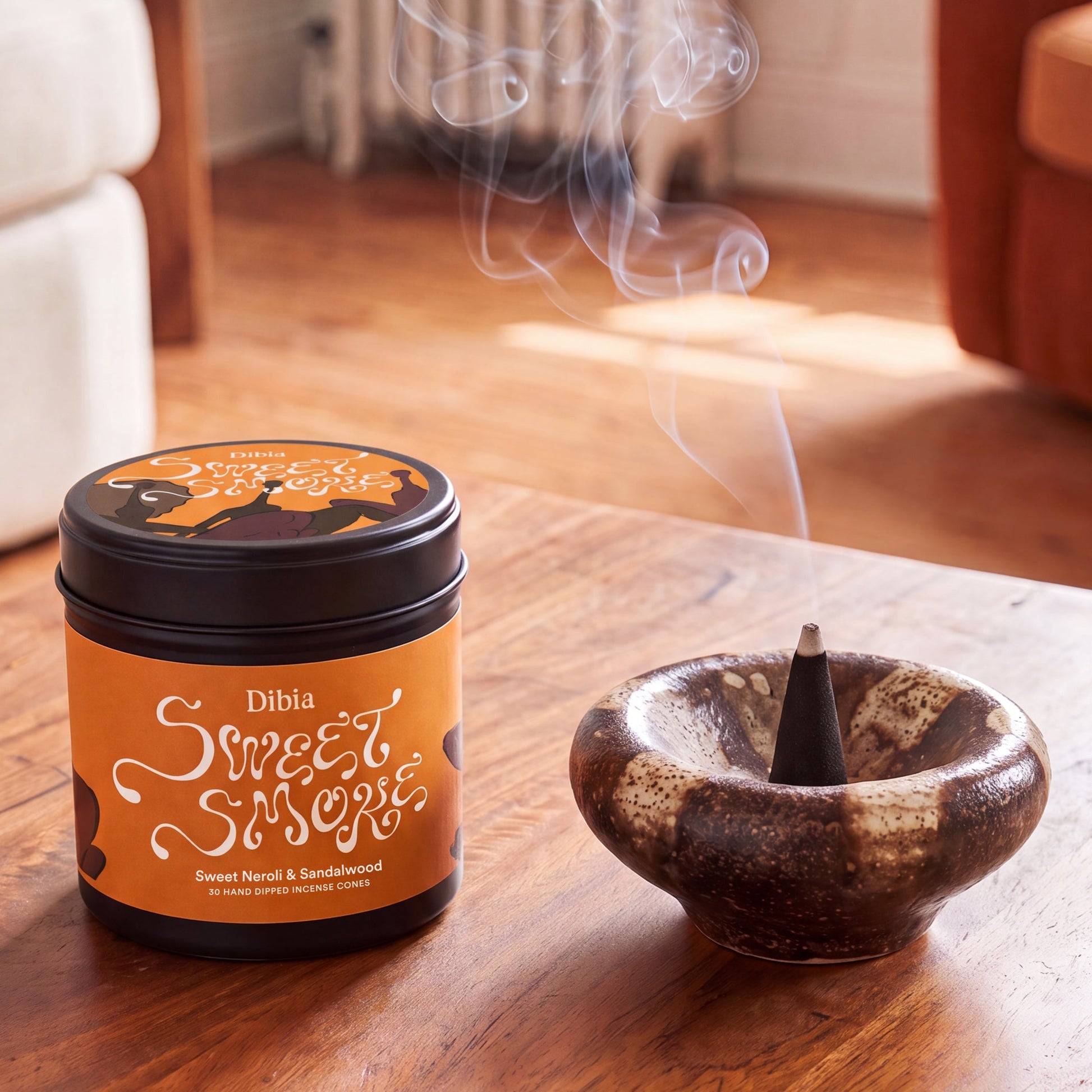 A jar of Dibia Sweet Smoky incense next to a ceramic incense holder with a smoking cone, set on a wooden table in a warm, softly blurred living room.