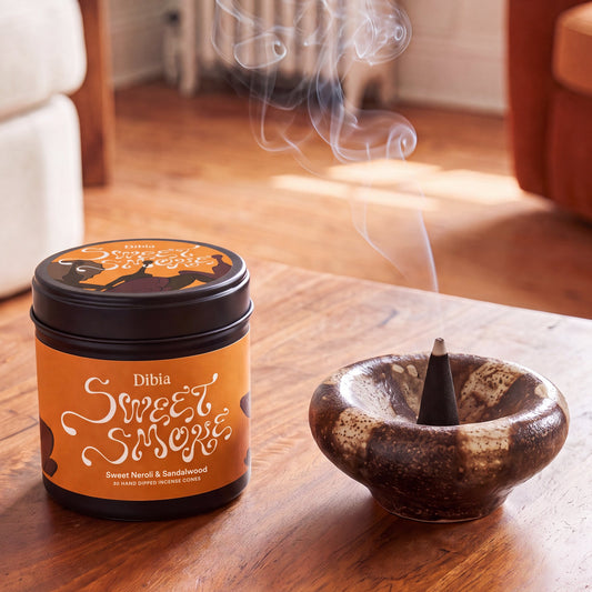 A jar of Dibia Sweet Smoky incense next to a ceramic incense holder with a smoking cone, set on a wooden table in a warm, softly blurred living room.
